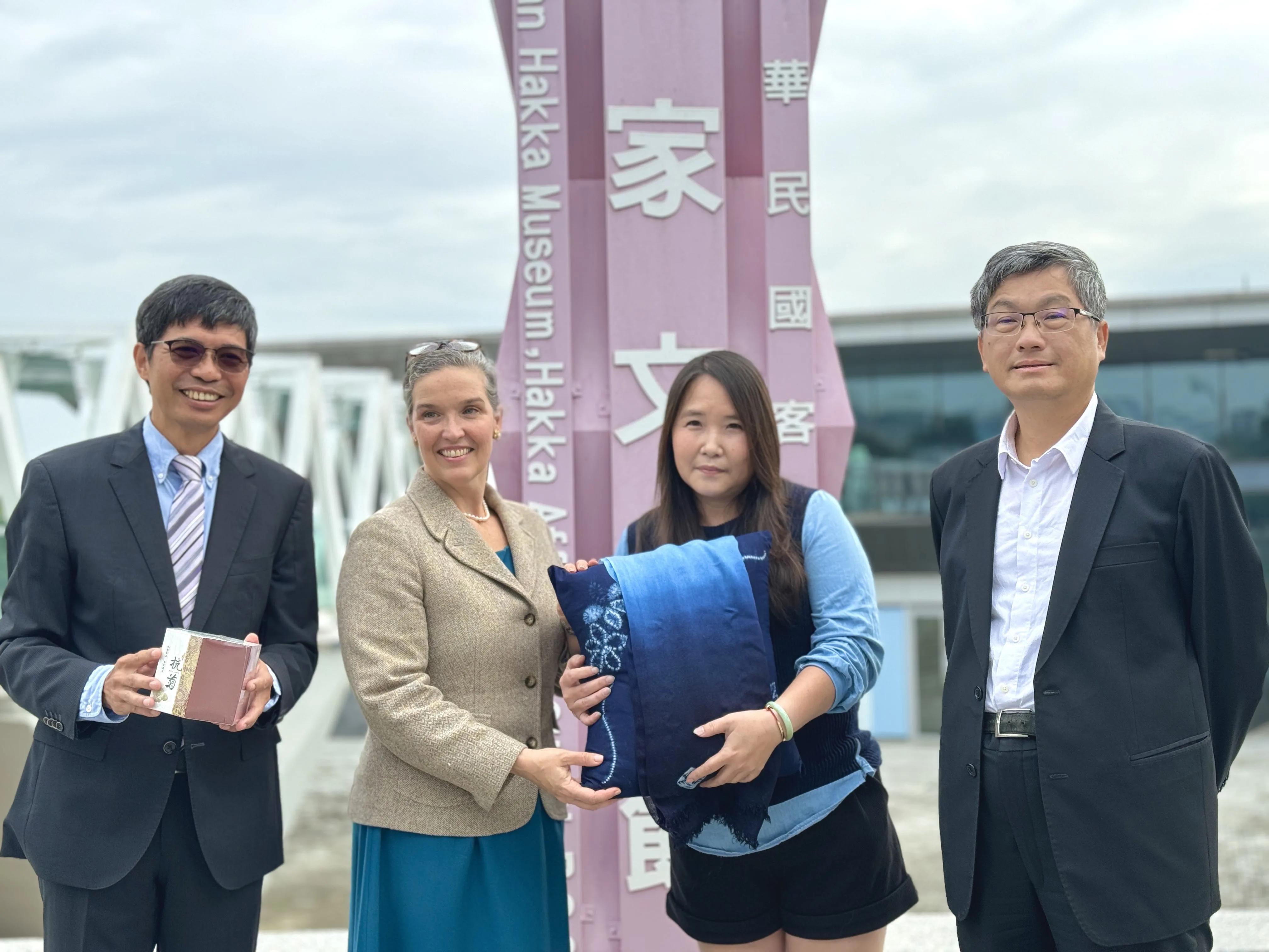 客委會主委楊長鎮(右一)、客發中心主任何金樑(左一)與教育推廣志工講師劉秀珍(右二)一起致贈客發中心環教藍染作品，歡迎AIT處長孫曉雅及其同仁前來臺灣客家文化館參訪。客發中心提供