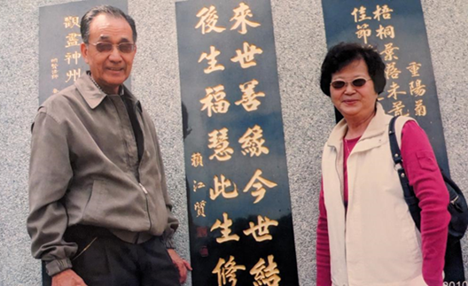 賴江椿夫婦回台到頭屋普光寺禮佛，在父親賴江質所書「來世善緣今世結，後生福慧此生修」詩牆前留影。賴江椿提供