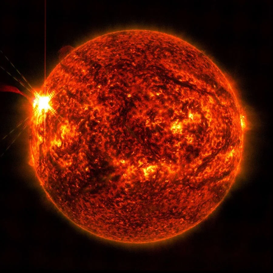 這張圖片由 NASA 的太陽動力學天文台於 5 月 14 日拍攝，顯示了一次太陽閃焰（左側明亮的閃光）。這張圖片呈現了一部分極紫外光，這種光線突顯了閃焰中極其高溫的物質，並以紅色著色。圖片來源：NASA/SDO